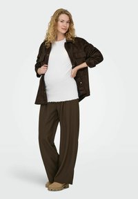 Brūna zamšādas jaka ar pogām pār baltu ribotu topu bez piedurknēm, kopā ar vaļīgām brūnām biksēm un bēšām kurpēm, ar vaļīgiem viļņainiem matiem.