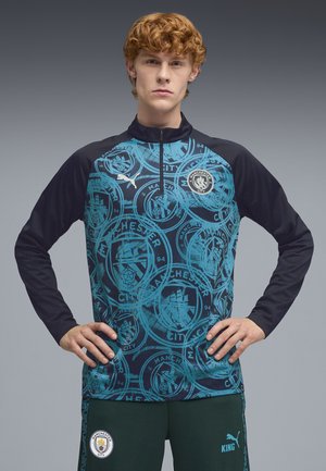Młody mężczyzna o kręconych rudych włosach stoi z rękami na biodrach, ubrany w niebiesko-czarną odzież sportową w wzory Manchester City z logo Puma.