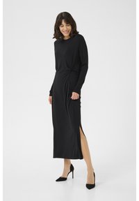 Black long-sleeve dress with a knotted waist detail, side slits, and a smooth texture, paired with black high-heeled shoes.