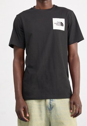 Homme portant un t-shirt noir à manches courtes avec un logo carré blanc "The North Face" sur le côté gauche de la poitrine et un jean bleu clair.
