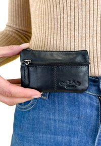 Černá kožená peněženka se zipovou kapsou, s prošívanými detaily a malou smyčkou pro upevnění, držena proti modrým džínům.