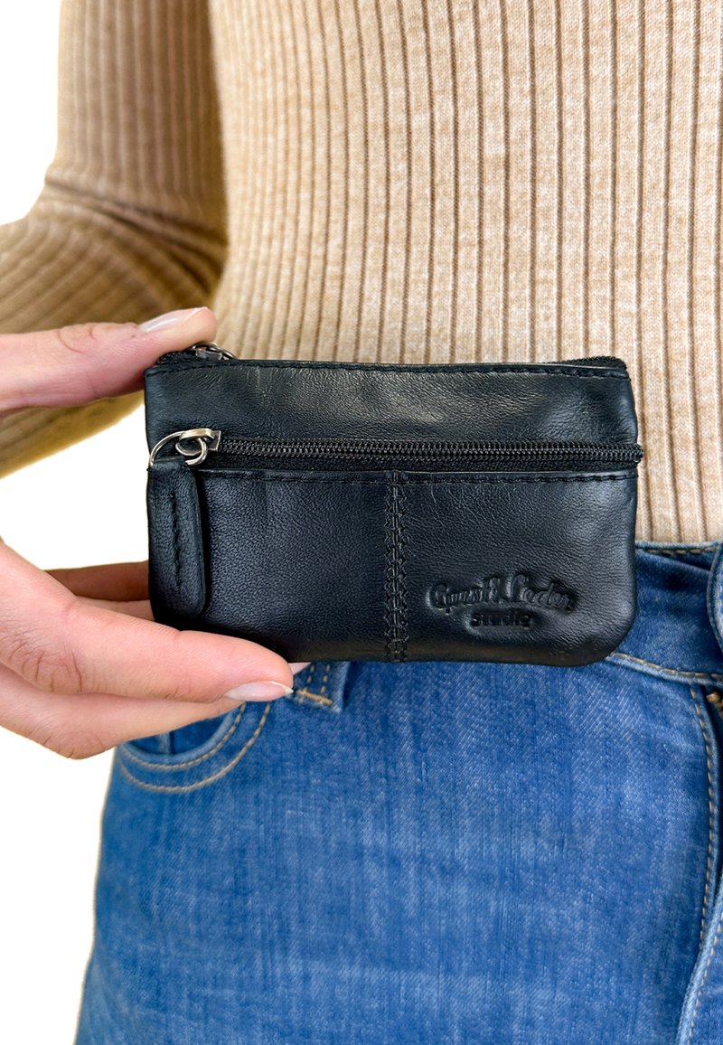 Černá kožená peněženka se zipovou kapsou, s prošívanými detaily a malou smyčkou pro upevnění, držena proti modrým džínům.