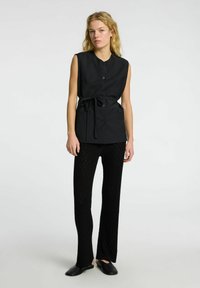 Top negro sin mangas con un escote redondo, botones frontales y un cintura atada. Combinado con pantalones negros de texturizados y zapatos negros de deslizar.