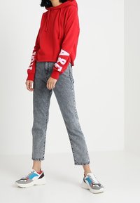 Felpa rossa con testo bianco sulle maniche, jeans grigi a vita alta e scarpe da ginnastica chunky bianche con accenti blu e rossi.