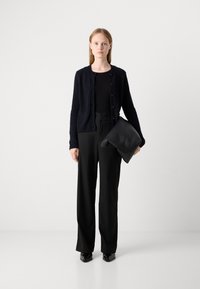 Frau in schwarzem strukturiertem Cardigan, schwarzem Top, weiten schwarzen Hosen und schwarzen Schuhen, die eine gefaltete schwarze Ledertasche vor einem schlichten Hintergrund hält.