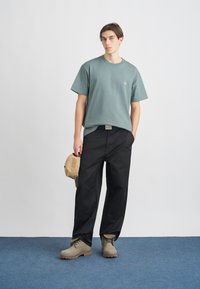 Lys grøn bomulds T-shirt med korte ærmer og et lille logo, parret med løse sorte bukser og beige snøresko. Brun kasket i hånden.