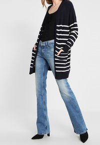 Mujer con un top negro con cordones, un cárdigan largo a rayas en tonos azul marino y blanco, unos vaqueros claros de pierna acampanada y unos tacones negros de punta.