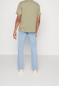 Olivgrön bomull t-shirt med korta ärmar, kombinerad med ljusblå raka jeans och mönstrade krämfärgade skor, sedd bakifrån.