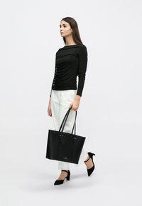 Μαύρο τοπ με μακριά μανίκια, πλαινό σούρωμα και λεπτομέρειες με κουμπιά, συνδυασμένο με λευκό παντελόνι και μεγάλη μαύρη τσάντα χειρός. Το μοντέλο φοράει μαύρες γόβες.