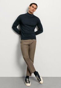 Navyfarbener Rollkragenpullover aus Strickstoff, kombiniert mit schlanken beigen Hosen. Getragen mit navyblauen Sneakers mit cremefarbenen Schnürsenkeln.