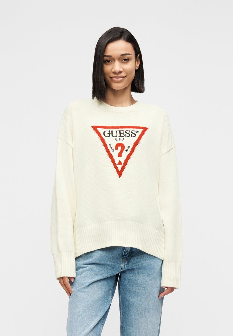 Γυναίκα με ίσια σκούρα μαλλιά φοράει λευκό oversized πουλόβερ με κόκκινο και μαύρο λογότυπο GUESS, συνδυασμένο με ανοιχτόχρωμο τζιν, σε απλό φόντο.