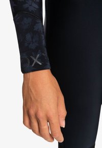Black wetsuit with floral pattern on the cuff, featuring a silver logo. Smooth, stretchy material highlights a fitted design.