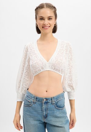 Jeune femme souriante, portant un crop top en dentelle blanche à manches longues et un jean bleu taille haute, sur un fond clair uni.