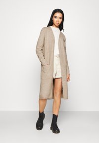 Cardigan lungo beige con una finitura testurizzata, apertura frontale e tasche laterali, indossato sopra una maglietta chiara e pantaloni corti beige, abbinato a stivali neri.