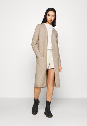 Cardigan lungo beige con una finitura testurizzata, apertura frontale e tasche laterali, indossato sopra una maglietta chiara e pantaloni corti beige, abbinato a stivali neri.