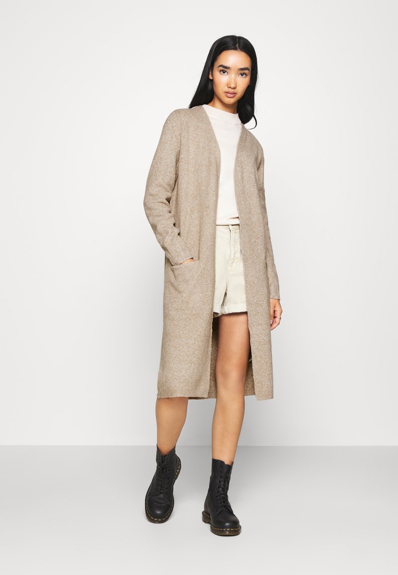 Cardigan lungo beige con una finitura testurizzata, apertura frontale e tasche laterali, indossato sopra una maglietta chiara e pantaloni corti beige, abbinato a stivali neri.