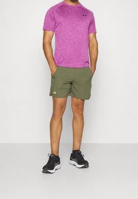 Muž oblečený v fialovém sportovním tričku s krátkým rukávem, olivově zelených šortkách s logem a černých běžeckých botách, stojí se založenýma rukama v kapsách.