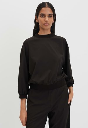 Donna con lunghi capelli scuri indossa una maglia nera a maniche lunghe larga e pantaloni neri abbinati su uno sfondo chiaro uniforme.
