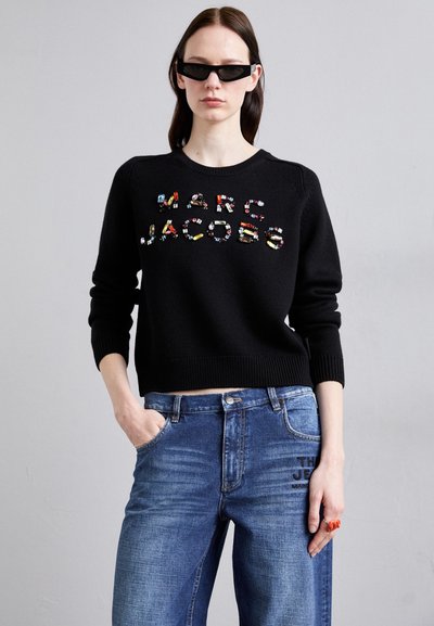Zwarte gebreide trui met kleurrijke geborduurde tekst "Marc Jacobs" aan de voorkant. Combineert met blauwe denim jeans met geprinte accenten.
