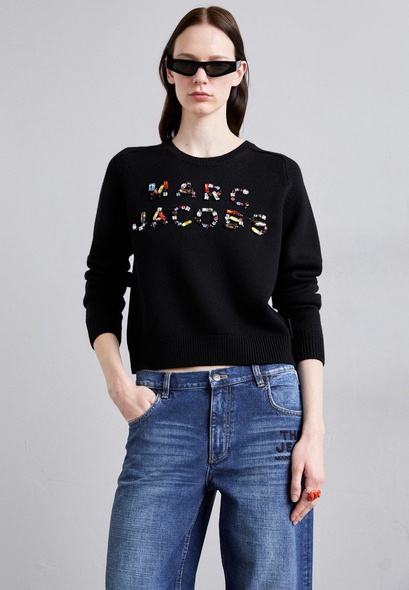 Pulover negru tricotat cu text brodat colorat "Marc Jacobs" pe față. Asortat cu blugi albaștri din denim cu accente imprimate.