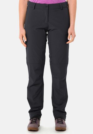 Frau trägt schwarze Wanderhose mit verstärkten Knien, steht aufrecht, kombiniert mit grauen und lila Wanderschuhen.