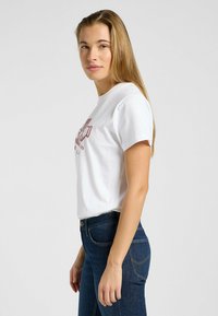 Camiseta de algodón blanca de manga corta, con un diseño gráfico rojo en la parte frontal. Combinada con jeans azul oscuro, que muestran una silueta ajustada.
