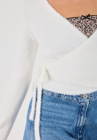 Weißes, flauschiges Wickeloberteil mit langen Ärmeln, das eine Schleife in der Taille hat, kombiniert mit hellblauen Jeans. Die untere Schicht hat ein Leopardenmuster.