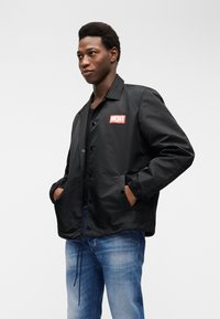 Νέος άνδρας που φοράει μαύρο μπουφάν με το λογότυπο Diesel και μπλε τζιν, με τα χέρια στις τσέπες, στέκεται μπροστά σε απλό λευκό φόντο.