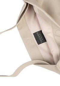 Beige tote bag met een gladde textuur, voorzien van een eenvoudig ontwerp, een binnenvak en een merklabel op een lichte stofvoering.