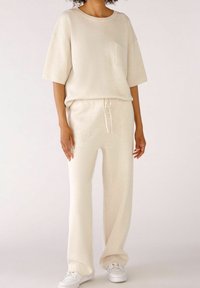 Osoba oblečená v krémovém krátkém rukávu s kapsou, v odpovídajících kalhotách se šňůrkou a bílých teniskách na světlém jednobarevném pozadí.