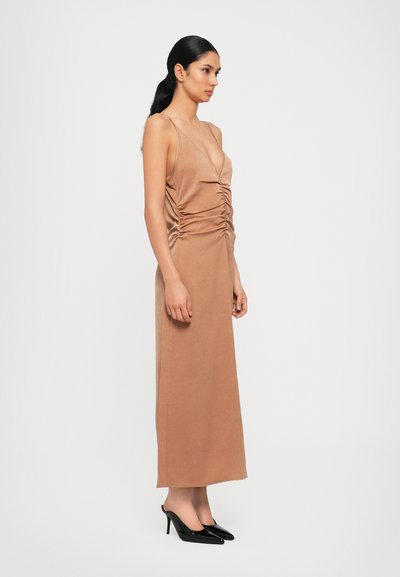 Long, body-hugging dress in a soft tan fabric with side ruching and a deep V-neckline, paired with black high-heeled shoes.