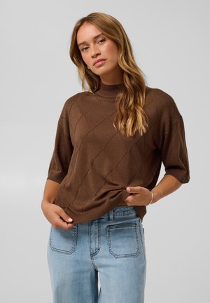 Jeune femme aux longs cheveux ondulés, portant un pull marron à manches courtes et un jean bleu clair, posant sur un fond clair uni.