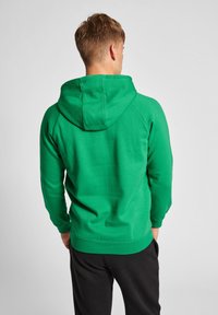 Grüner Pullover-Hoodie mit Kordelzugkapuze, gerippten Bündchen und Saum, mit einer glatten Textur und einem schlichten Design. Schwarze Hosen sind darunter sichtbar.