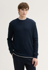 Maglione a coste blu navy con maniche lunghe e scollatura rotonda, con un accento bianco all'orlo. Indossato sopra una maglietta bianca.