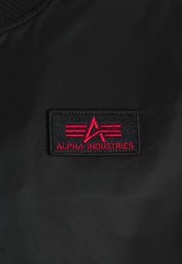 Zwarte bomberjackstof met een rood geborduurd patch waarop de tekst "ALPHA INDUSTRIES" en grafische elementen staan, gelegen op het borstgedeelte.