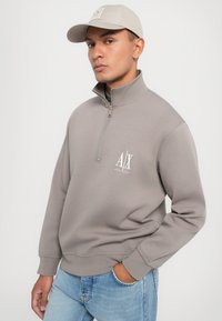 Sudadera gris de cuello alto con media cremallera, textura suave, puños acanalados y un logo blanco en el pecho. Combinada con jeans de mezclilla azul.