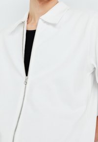 Veste blanche avec col et fermeture éclair centrale. Confectionnée en tissu lisse, avec une coupe ajustée et un design minimaliste.