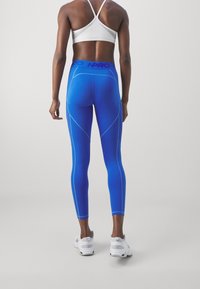 Blaue Leggings mit kontrastierenden hellblauen Nähten, hohem Bündchen und einem figurbetonten Design. Kombiniert mit einem weißen Sport-BH und weißen Turnschuhen.