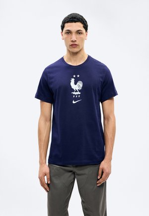 Noor mees seisab näoga otse, kandes tumesinist T-särki, millel on valge kukelogo, kaks tähte, tähed "FFF" ja Nike logo.