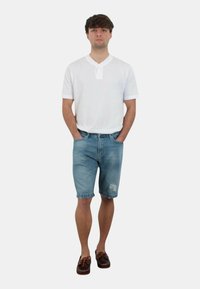 Maglietta henley a maniche corte bianca, pantaloni corti di jeans azzurri chiari con lievi segni di usura e scarpe da barca marroni. Il modello sta in piedi con le mani in tasca.