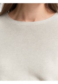 Gros plan d'une personne portant un haut côtelé beige clair à col rond, montrant le décolleté et le haut de la poitrine avec de longs cheveux bruns visibles.