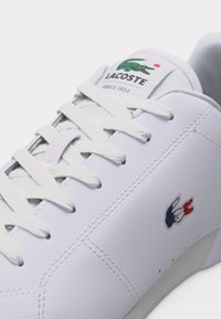 Nærbilde av hvit Lacoste-sneakers med teksturerte lisser, detaljer i sømmen og brodert rød, hvit og blå krokodillelogo på siden.
