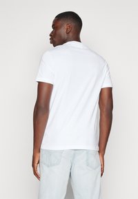 Hvit bomulls-t-skjorte med korte ermer og ribbestrikket krage, vist fra baksiden. Et par lyseblå jeans med baklommer delvis synlige.