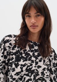 Tânără cu părul închis la culoare, până la umeri, purtând un top alb și negru cu un model abstract, pe un fundal deschis simplu.