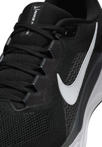 Chaussure de sport noire avec une tige en mesh, dotée du logo Swoosh blanc de Nike, de lacets texturés et d'une semelle rembourrée.