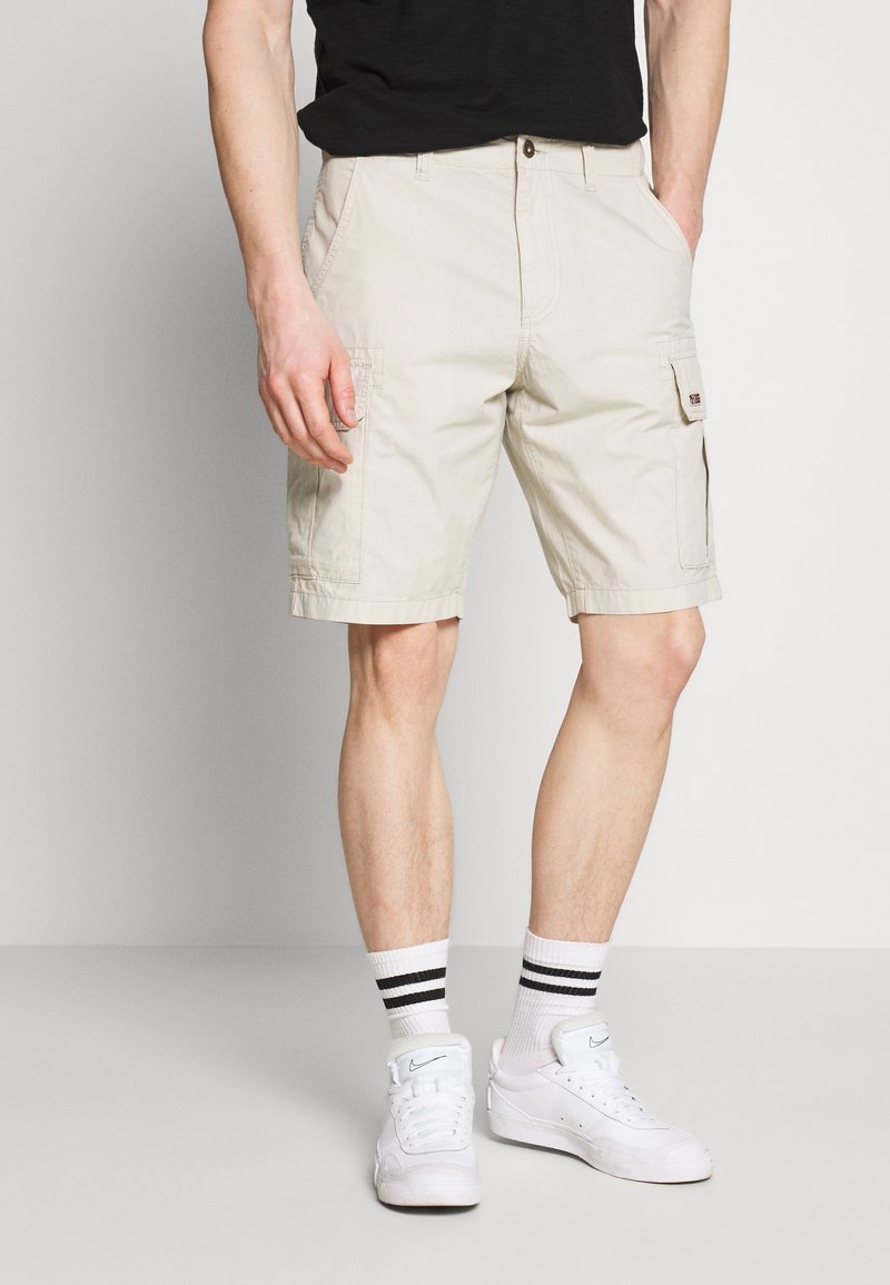 Shorts cargo beiges avec poches avant et latérales, fabriqués en coton. Portés avec des baskets blanches et des chaussettes rayées. Design simple, longueur jusqu'au genou.