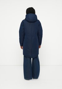 Cappotto lungo imbottito blu navy con cappuccio, con vita cinchata, tasche laterali e orlo arrotondato. Indossato sopra pantaloni in denim a gamba larga.