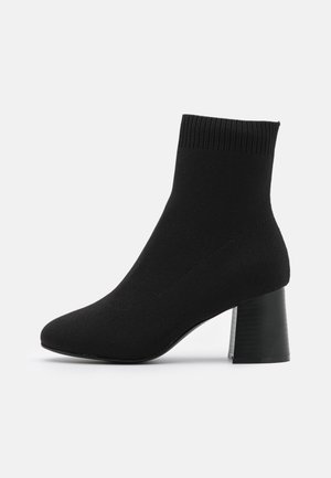 Schwarzer Knöchelsocken-Stiefel mit geripptem Bund und Blockabsatz, Seitenansicht auf weißem Hintergrund.