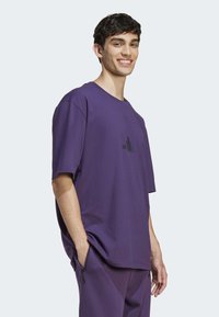 Violet trumpas rankovėmis marškinėliai, pagaminti iš medvilnės, su apvaliu iškirpimu ir juodu logotipu, atspausdintu priekyje. Atsitiktinis, laisvas kirpimas.