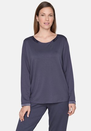 Donna in piedi che indossa una maglia grigio scuro a maniche lunghe e pantaloni coordinati con uno sfondo chiaro semplice.